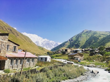 A picturesque mountain village with traditional stone houses under a clear blue sky. A river flows through the area, surrounded by lush green hills and snow-capped peaks in the distance.