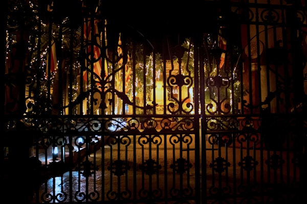 An ornate black iron gate is silhouetted against a backdrop of warm yellow and orange lights. Behind the gate, decorative lights wrap around trees and plants, adding a festive atmosphere to the scene. The ground is wet, reflecting some of the lights, giving the image an enchanting and mysterious quality.