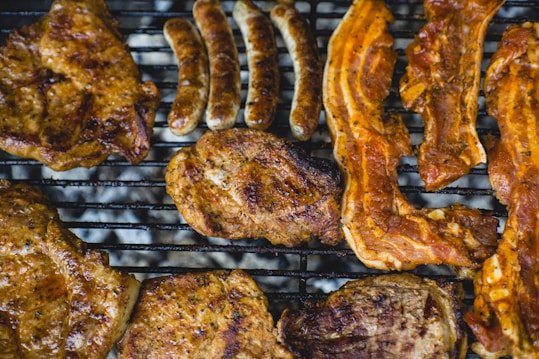 A variety of meats such as sausages, pork chops, and bacon strips are placed on a grill. The meat appears to be seasoned and cooked, with grill marks visible across the surface. The scene evokes a casual barbecue setting with a close-up view emphasizing the texture and color of the grilled meats.