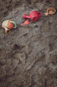 A sandy surface with scattered children's beach toys, including colorful plastic watering cans and a small toy in the shape of a fish or foot.