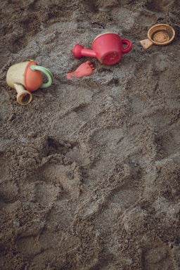 A sandy surface with scattered children's beach toys, including colorful plastic watering cans and a small toy in the shape of a fish or foot.