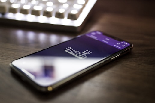 A smartphone is placed on a desk, displaying the Twitch logo on its screen. The device is positioned at an angle, and there is a blurred computer keyboard in the background, suggesting a desk setup related to streaming or gaming.