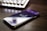 A smartphone is placed on a desk, displaying the Twitch logo on its screen. The device is positioned at an angle, and there is a blurred computer keyboard in the background, suggesting a desk setup related to streaming or gaming.