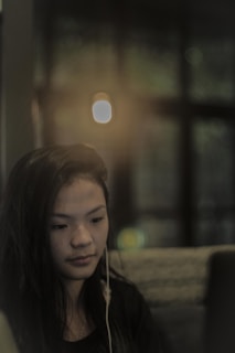 A woman with long dark hair is wearing earphones while looking at a laptop screen. The setting is dimly lit, with a blurred background showing a soft focus light, suggesting an indoor and cozy environment.