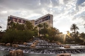 A luxurious hotel with prominent signage is surrounded by lush greenery and palm trees. In the foreground, a series of tiered waterfalls and rock formations add a natural element. The sun shines through the clouds, creating a warm and inviting atmosphere.
