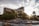 A luxurious hotel with prominent signage is surrounded by lush greenery and palm trees. In the foreground, a series of tiered waterfalls and rock formations add a natural element. The sun shines through the clouds, creating a warm and inviting atmosphere.