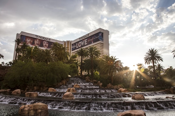 A luxurious hotel with prominent signage is surrounded by lush greenery and palm trees. In the foreground, a series of tiered waterfalls and rock formations add a natural element. The sun shines through the clouds, creating a warm and inviting atmosphere.