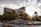 A luxurious hotel with prominent signage is surrounded by lush greenery and palm trees. In the foreground, a series of tiered waterfalls and rock formations add a natural element. The sun shines through the clouds, creating a warm and inviting atmosphere.