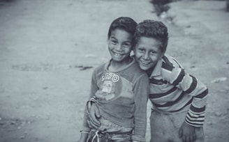 Two young boys are standing close together, smiling warmly at the camera. The boy on the left is wearing a long-sleeved shirt with a graphic print, while the boy on the right is dressed in a striped shirt. The background is a paved area, slightly blurred, emphasizing the joyful expressions of the boys.