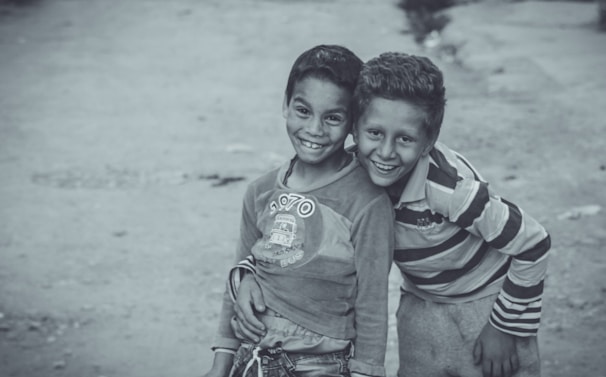 Two young boys are standing close together, smiling warmly at the camera. The boy on the left is wearing a long-sleeved shirt with a graphic print, while the boy on the right is dressed in a striped shirt. The background is a paved area, slightly blurred, emphasizing the joyful expressions of the boys.