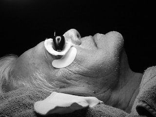 A black and white image of a person lying down with their head slightly tilted back. The individual has a device or contraption around their nose, possibly for medical or therapeutic purposes. The person's hair appears to be gray or white, and their skin shows visible texture and minor stubble. There is a towel or cloth draped around their neck and shoulders.