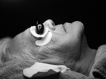 A black and white image of a person lying down with their head slightly tilted back. The individual has a device or contraption around their nose, possibly for medical or therapeutic purposes. The person's hair appears to be gray or white, and their skin shows visible texture and minor stubble. There is a towel or cloth draped around their neck and shoulders.
