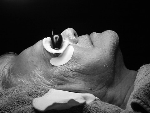 A black and white image of a person lying down with their head slightly tilted back. The individual has a device or contraption around their nose, possibly for medical or therapeutic purposes. The person's hair appears to be gray or white, and their skin shows visible texture and minor stubble. There is a towel or cloth draped around their neck and shoulders.