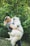 A person smiling while holding a large fluffy white dog in their arms. They are standing in a lush green forest with dense foliage. The dog appears happy and relaxed.