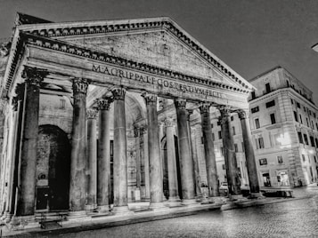 A grayscale image of an ancient, monumental structure with large Corinthian columns and a triangular pediment. The inscription on the pediment indicates historical significance. Surrounding buildings show classic architectural designs, and the cobblestone pavement adds texture to the scene.