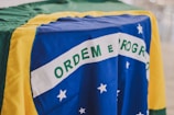 A close-up of the Brazilian flag displaying the green, yellow, and blue sections with white stars and the phrase 'Ordem e Progresso' prominently. The fabric appears to be slightly wrinkled, indicating it is draped over a surface.