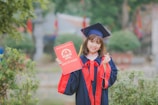 woman wearing black and red academic dress and mortar board holding red book cover near green grass