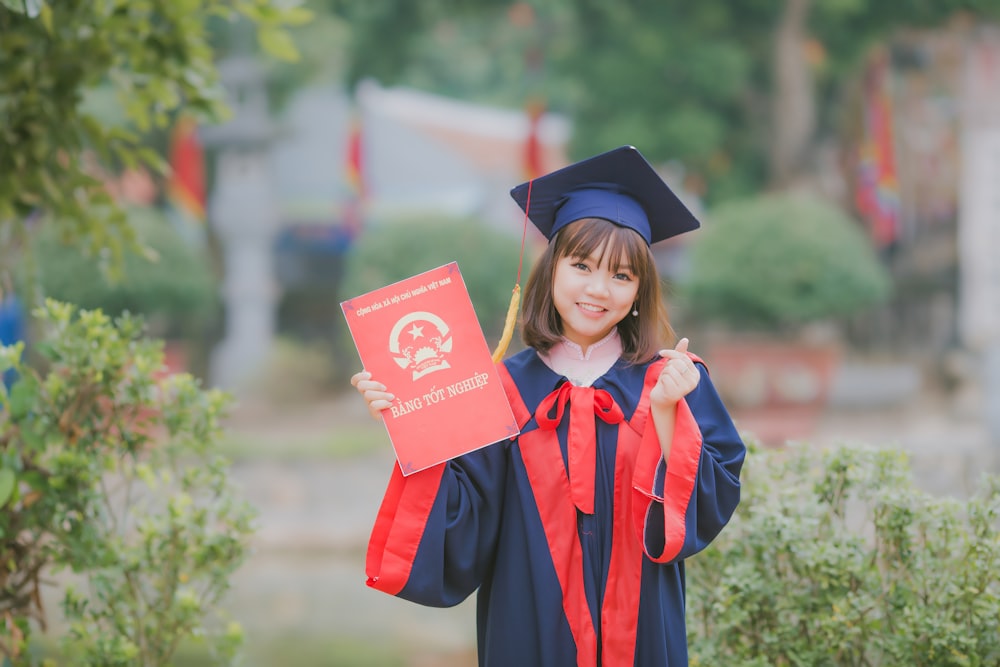 woman wearing black and red academic dress and mortar board holding red book cover near green grass