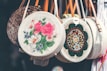 Circular woven handbags hang in a row, each with unique and colorful embroidery. The central bag features bold pink and white floral patterns, while another displays intricate geometric designs. Leather straps add a touch of contrast to the baskets.