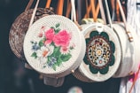 Circular woven handbags hang in a row, each with unique and colorful embroidery. The central bag features bold pink and white floral patterns, while another displays intricate geometric designs. Leather straps add a touch of contrast to the baskets.