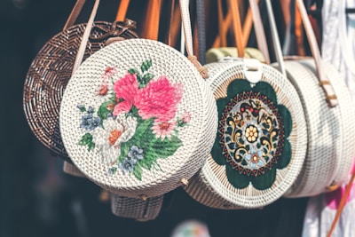 Circular woven handbags hang in a row, each with unique and colorful embroidery. The central bag features bold pink and white floral patterns, while another displays intricate geometric designs. Leather straps add a touch of contrast to the baskets.