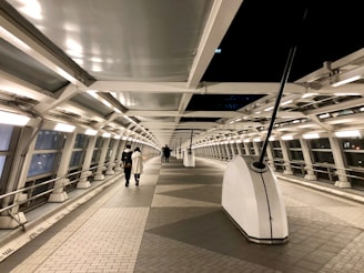 A modern pedestrian bridge with a long, curved corridor enclosed by glass and metal beams. The walkway is illuminated by evenly spaced lights along the sides, creating a calm and futuristic atmosphere. Two people are walking toward the far end of the bridge.