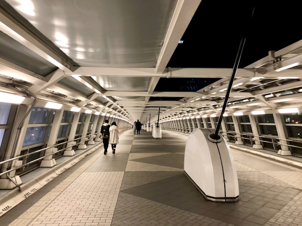 A modern pedestrian bridge with a long, curved corridor enclosed by glass and metal beams. The walkway is illuminated by evenly spaced lights along the sides, creating a calm and futuristic atmosphere. Two people are walking toward the far end of the bridge.
