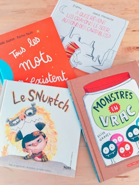 Four colorful children's books are arranged on a light wooden surface. Each book cover features distinct artwork and typography. One book has an orange cover with white lettering and small star designs. Another book has a blue and white cover with an illustration of an indigenous character. A third book displays an animated character with a furry costume on a primarily white cover. The fourth book has a cartoonish jar filled with bright-colored characters against a brownish background.