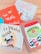 Four colorful children's books are arranged on a light wooden surface. Each book cover features distinct artwork and typography. One book has an orange cover with white lettering and small star designs. Another book has a blue and white cover with an illustration of an indigenous character. A third book displays an animated character with a furry costume on a primarily white cover. The fourth book has a cartoonish jar filled with bright-colored characters against a brownish background.