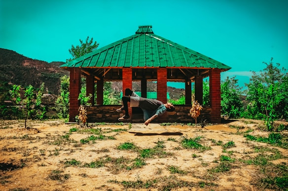 A person is performing a backbend or a yoga pose in front of a small open pavilion with a green metal roof. The structure has brick pillars and is surrounded by lush greenery and hills in the background under a bright blue sky.