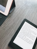 A wooden surface with a tablet and an e-reader. The tablet is in a black case, partly visible on the left. The e-reader displays text in French, positioned diagonally on the right side.