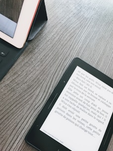 A wooden surface with a tablet and an e-reader. The tablet is in a black case, partly visible on the left. The e-reader displays text in French, positioned diagonally on the right side.