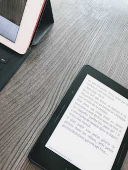 A wooden surface with a tablet and an e-reader. The tablet is in a black case, partly visible on the left. The e-reader displays text in French, positioned diagonally on the right side.