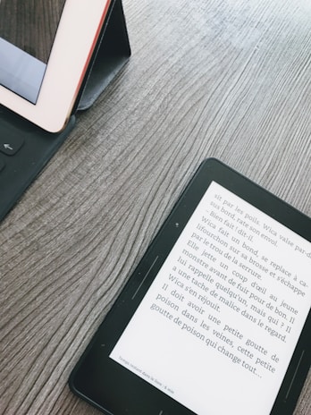 A wooden surface with a tablet and an e-reader. The tablet is in a black case, partly visible on the left. The e-reader displays text in French, positioned diagonally on the right side.