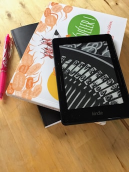 A Kindle device displaying a black and white image of a typewriter's keys is placed on top of a stack of two books with colorful covers. Next to the books is a pink pen, all resting on a wooden surface.