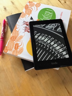 A Kindle device displaying a black and white image of a typewriter's keys is placed on top of a stack of two books with colorful covers. Next to the books is a pink pen, all resting on a wooden surface.