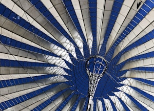 A modern architectural structure with a radial, tent-like design that includes metal beams and a fabric material. The structure forms a circular pattern with alternating blue and white segments. The blue sections are likely open spaces or covered with transparent material, giving a view of the clear sky above.