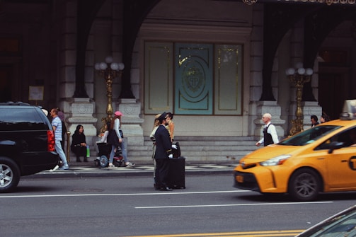 A busy urban street scene featuring several people walking and interacting near the entrance of an ornate building with large doors and decorative elements. A bellman in uniform stands with a suitcase on the sidewalk. A yellow taxi is in motion on the street, and there is a black SUV parked nearby. A few people are seated near the entrance, some standing or walking, and one person is riding a scooter.