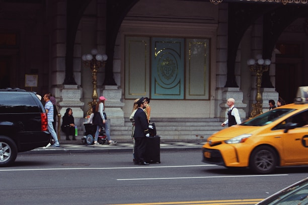 A busy urban street scene featuring several people walking and interacting near the entrance of an ornate building with large doors and decorative elements. A bellman in uniform stands with a suitcase on the sidewalk. A yellow taxi is in motion on the street, and there is a black SUV parked nearby. A few people are seated near the entrance, some standing or walking, and one person is riding a scooter.