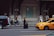 A busy urban street scene featuring several people walking and interacting near the entrance of an ornate building with large doors and decorative elements. A bellman in uniform stands with a suitcase on the sidewalk. A yellow taxi is in motion on the street, and there is a black SUV parked nearby. A few people are seated near the entrance, some standing or walking, and one person is riding a scooter.