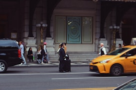 A busy urban street scene featuring several people walking and interacting near the entrance of an ornate building with large doors and decorative elements. A bellman in uniform stands with a suitcase on the sidewalk. A yellow taxi is in motion on the street, and there is a black SUV parked nearby. A few people are seated near the entrance, some standing or walking, and one person is riding a scooter.