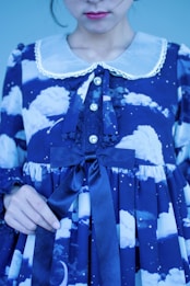 A person is wearing a blue dress with a pattern of clouds, stars, and a crescent moon. The dress features a large, dark ribbon tied into a bow at the waist. The collar is white with lace trim, and the dress has decorative buttons.