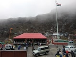 A large group of people is gathered around a building with a red roof located in a mountainous area. There is a prominent flagpole displaying the Indian national flag. Several parked vehicles, including cars and an SUV, are visible. The background consists of fog-covered hills and rocky terrain.