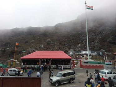 A large group of people is gathered around a building with a red roof located in a mountainous area. There is a prominent flagpole displaying the Indian national flag. Several parked vehicles, including cars and an SUV, are visible. The background consists of fog-covered hills and rocky terrain.
