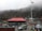 A large group of people is gathered around a building with a red roof located in a mountainous area. There is a prominent flagpole displaying the Indian national flag. Several parked vehicles, including cars and an SUV, are visible. The background consists of fog-covered hills and rocky terrain.