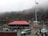 A large group of people is gathered around a building with a red roof located in a mountainous area. There is a prominent flagpole displaying the Indian national flag. Several parked vehicles, including cars and an SUV, are visible. The background consists of fog-covered hills and rocky terrain.