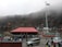 A large group of people is gathered around a building with a red roof located in a mountainous area. There is a prominent flagpole displaying the Indian national flag. Several parked vehicles, including cars and an SUV, are visible. The background consists of fog-covered hills and rocky terrain.
