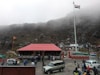 A large group of people is gathered around a building with a red roof located in a mountainous area. There is a prominent flagpole displaying the Indian national flag. Several parked vehicles, including cars and an SUV, are visible. The background consists of fog-covered hills and rocky terrain.