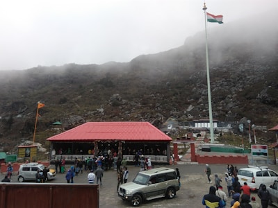 A large group of people is gathered around a building with a red roof located in a mountainous area. There is a prominent flagpole displaying the Indian national flag. Several parked vehicles, including cars and an SUV, are visible. The background consists of fog-covered hills and rocky terrain.