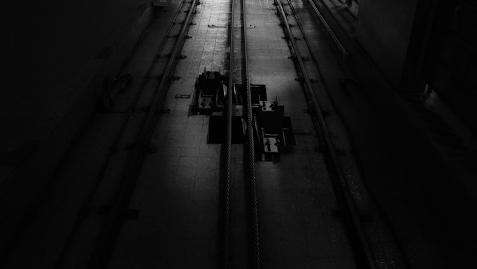 A dimly lit industrial setting with two cables running vertically along a textured floor. There are mechanical components in the center, casting shadows due to the minimal lighting.
